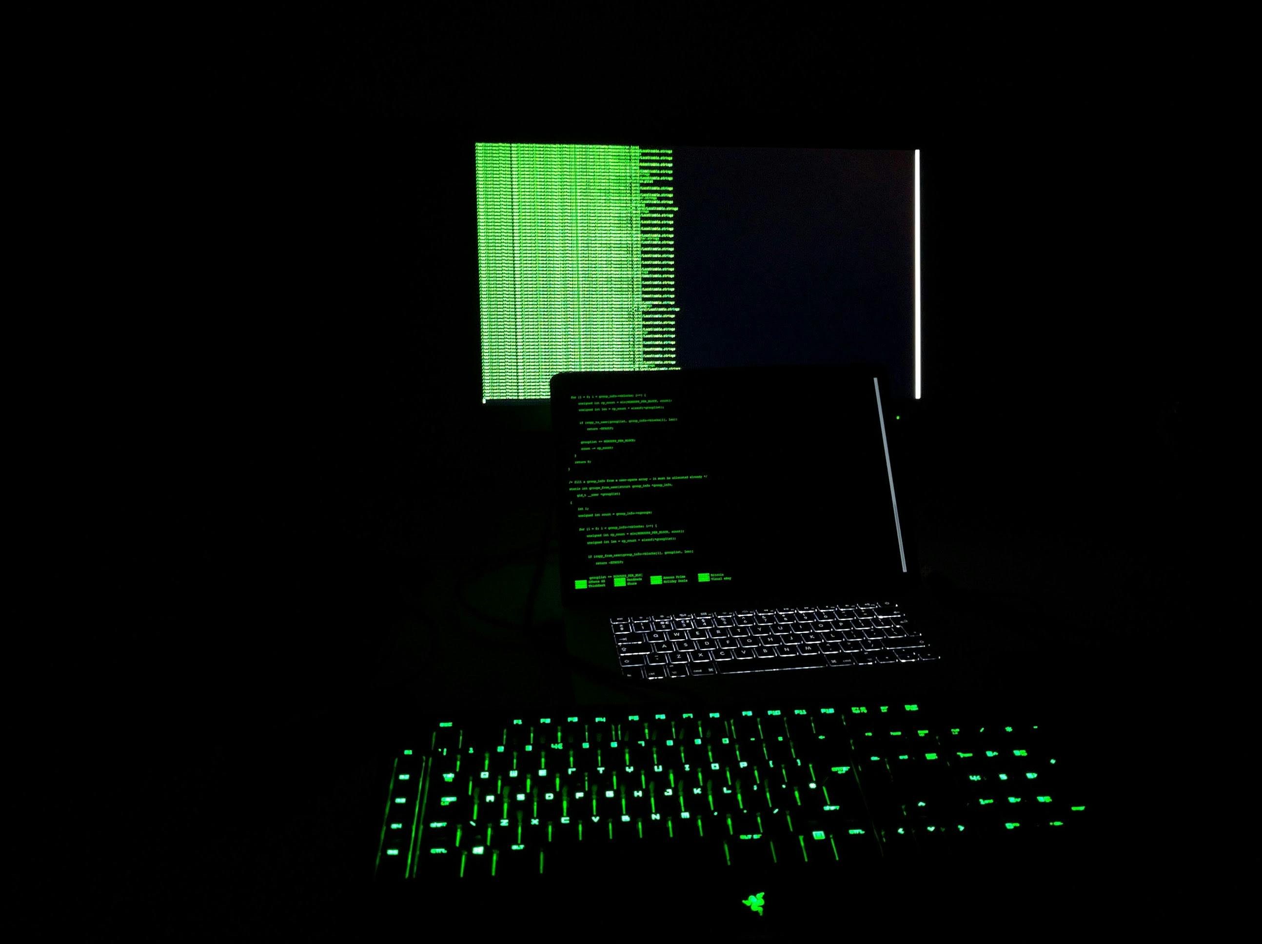 A dark room illuminated by the green glow of a backlit mechanical keyboard in the foreground and a laptop screen displaying lines of code, with a secondary monitor showing similar content in the background.